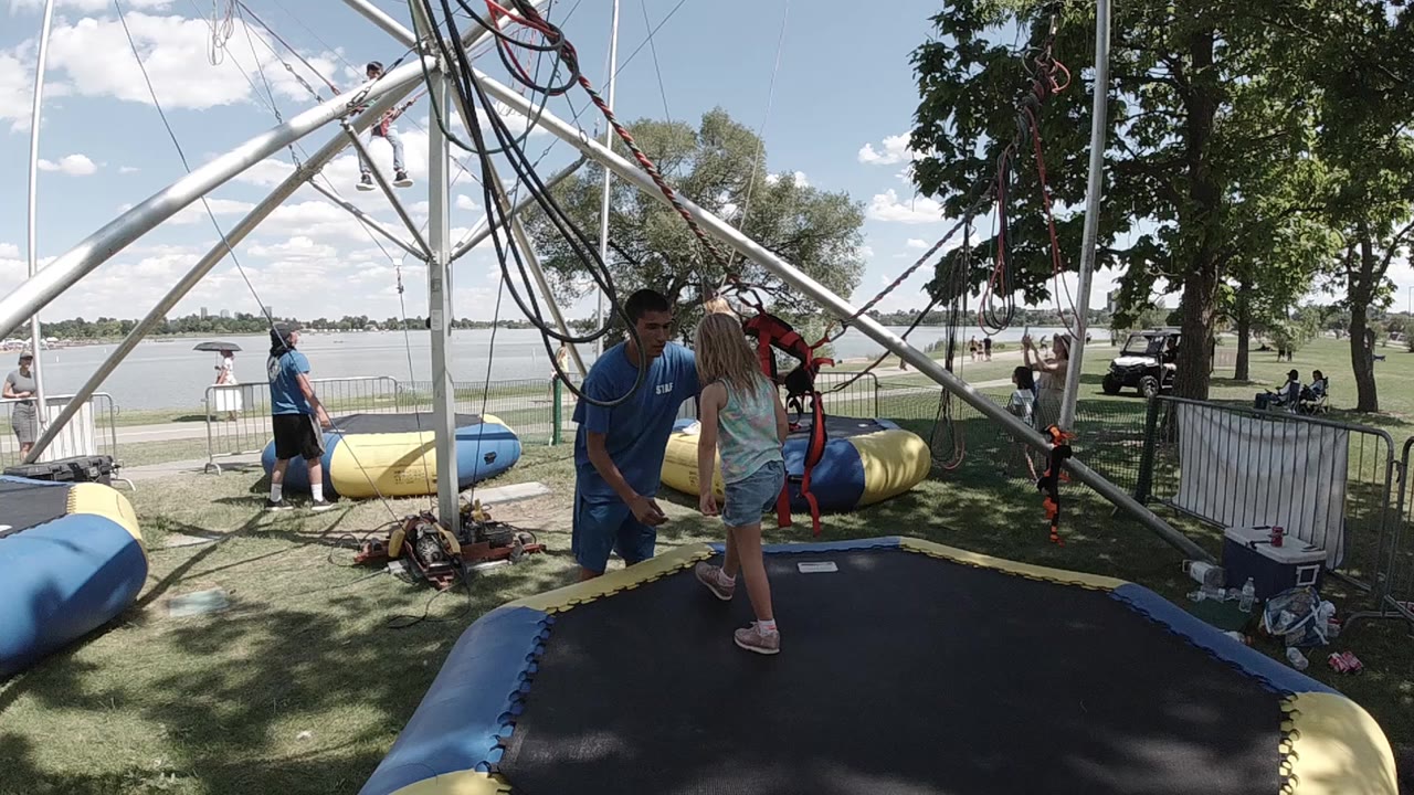 At a Colorado outdoor attraction, a blue-shirted instructor stands near a child on a black trampoline mat beneath bungee cords and metal supports.