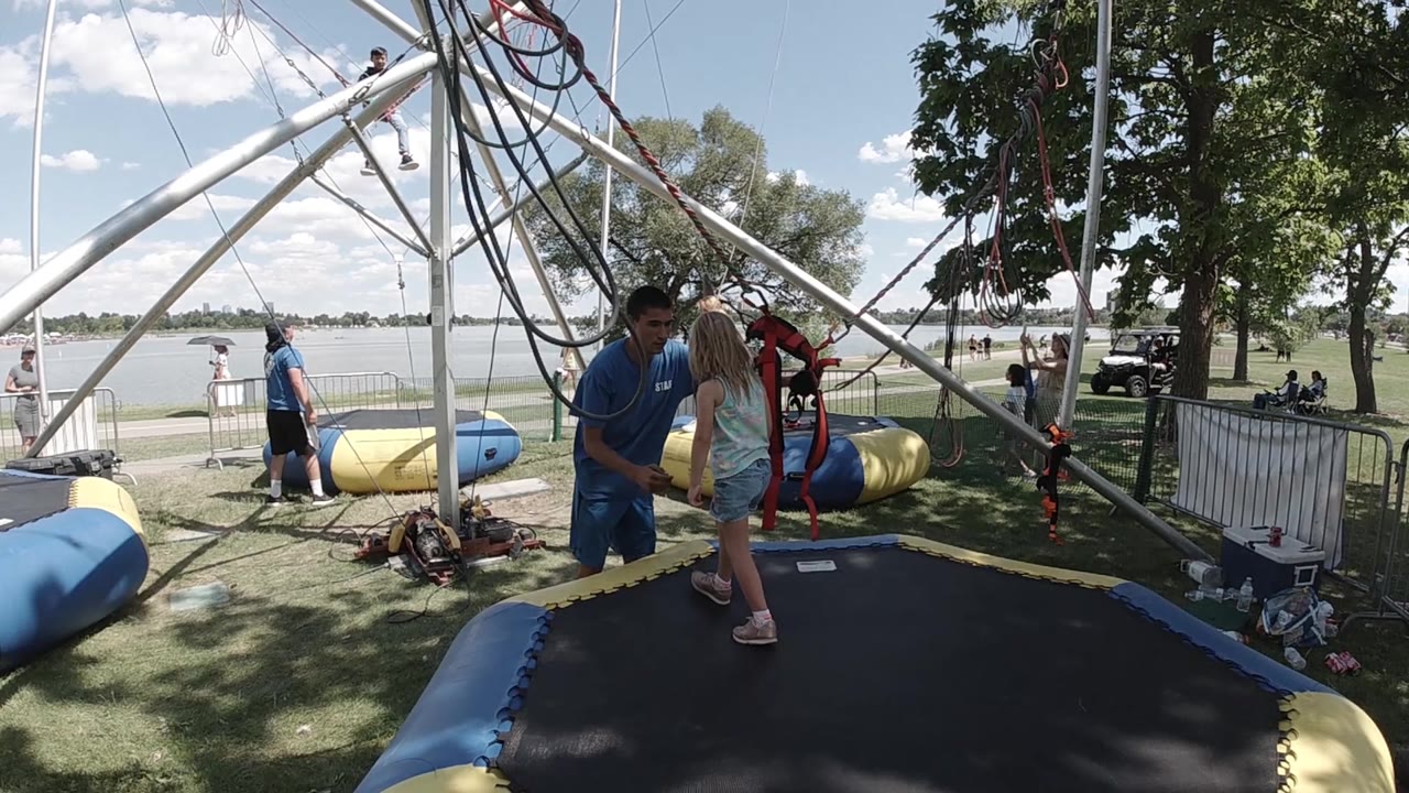 The instructor faces the child at the edge of the trampoline area, with red harness straps visible in front of them and the bungee rig overhead.