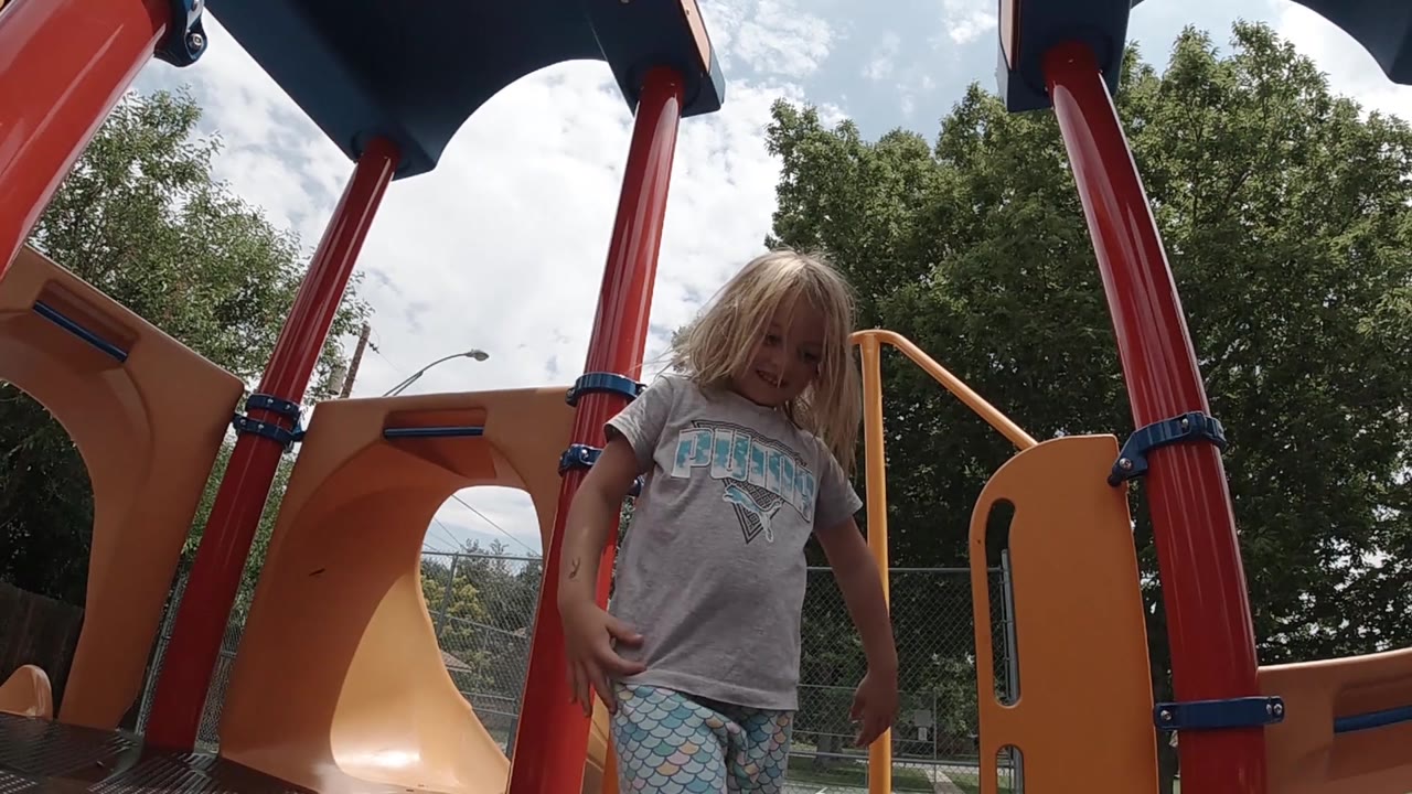 The child shifts slightly beside a red support pole and orange railing, with trees, sky, and playground hardware visible around her.