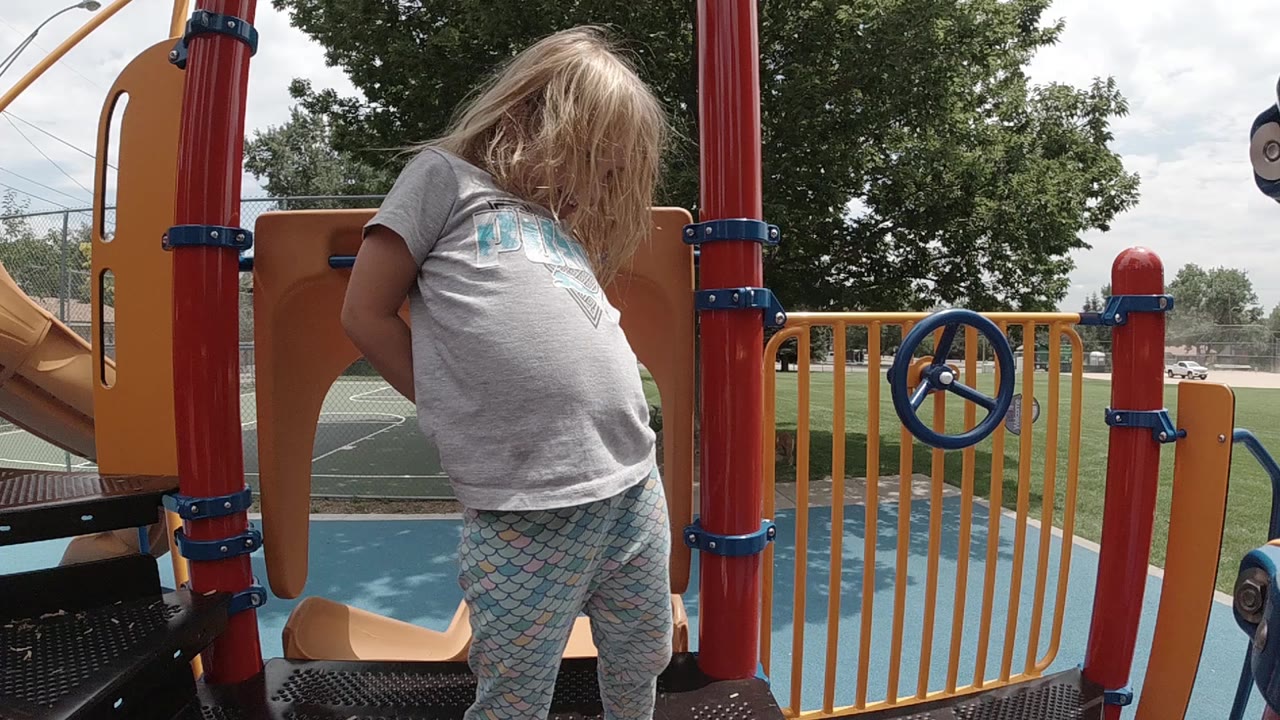 A blonde toddler in a gray shirt stands on a Colorado playground ship deck between red support posts, orange panels, yellow railings, and a blue steering wheel.