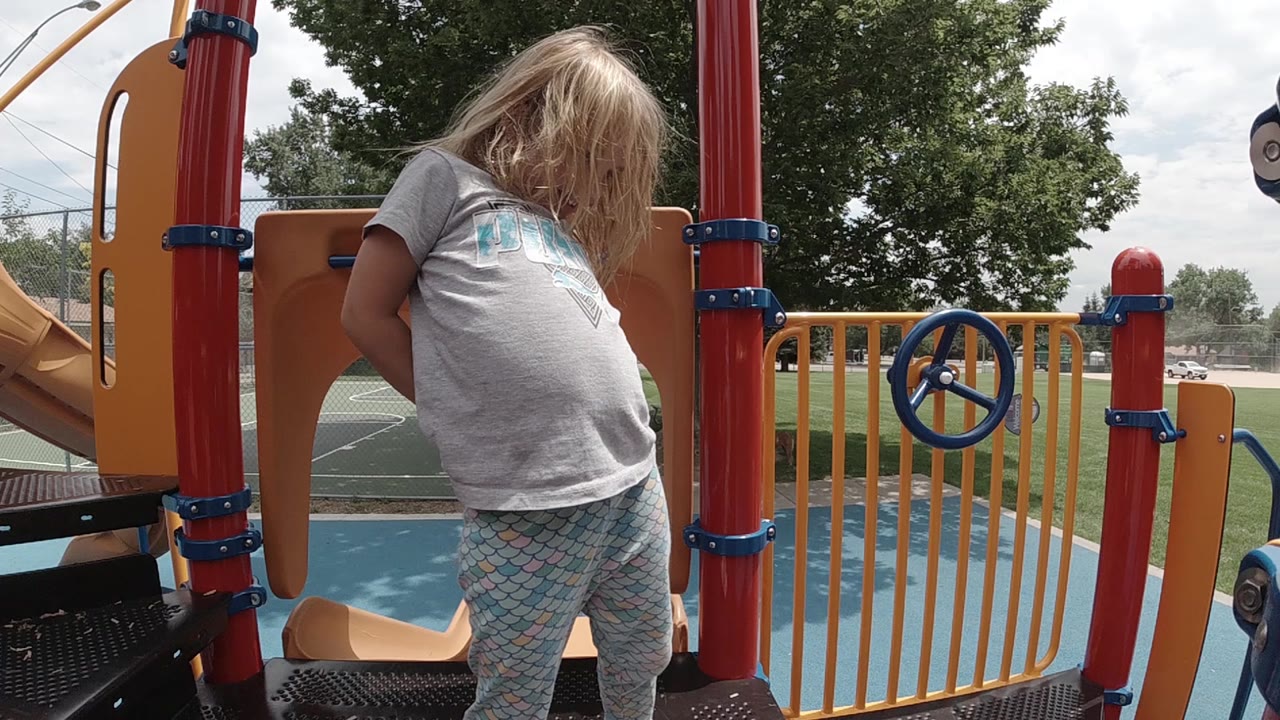 Toddler leans slightly on the playground deck near the yellow railing and blue wheel.