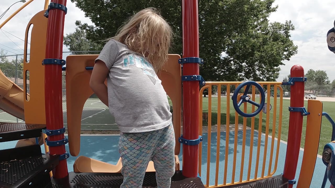 A blonde child stands on the Colorado playground platform with hair falling forward, framed by red posts, orange panels, yellow railings, and park trees behind.