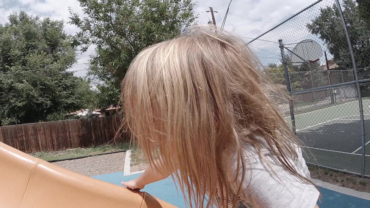 In Colorado, a blonde young girl leans over the side of a tan plastic playground slide with trees, chain-link fencing, and a basketball hoop in the background.