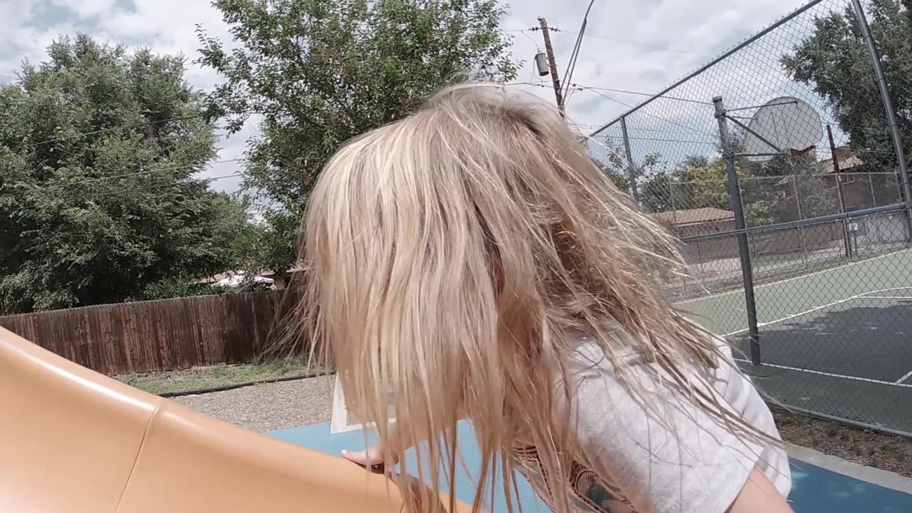 The girl bends forward beside the slide, hair falling across her face as the outdoor fence, trees, and hoop frame the residential background.