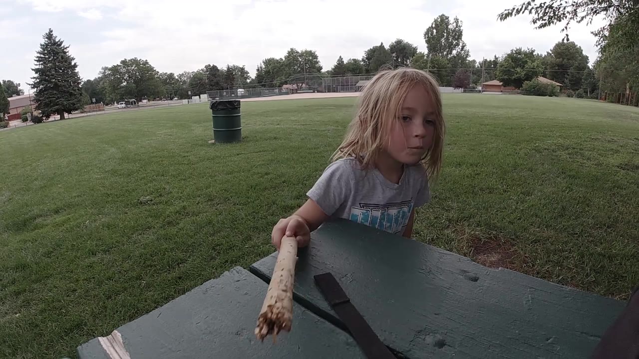 The child brings the muddy end of the wooden stick toward the green picnic table, with the black strap lying across the tabletop.