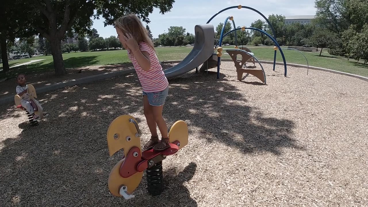 At 0:00 in a Colorado playground, a toddler girl in pink stands over a yellow bull-shaped spring rider while another child sits near the left edge and a slide is visible behind her.