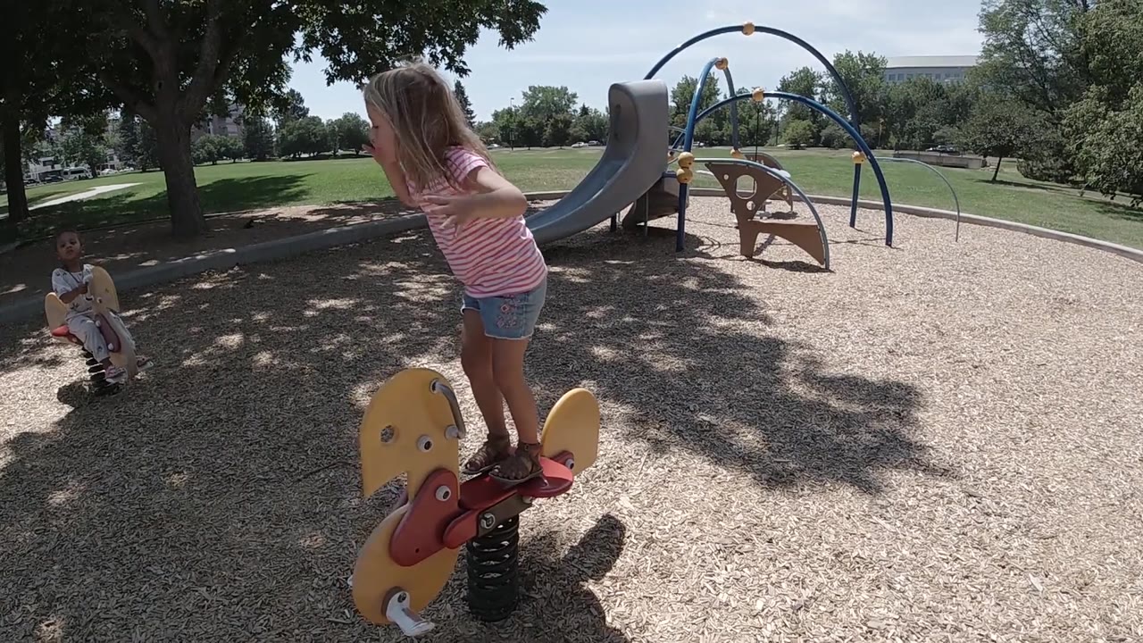 At 0:02, the toddler bends forward over the yellow bull-shaped rider as the sunny playground, slide, climbing structure, and shaded trees remain in the background.
