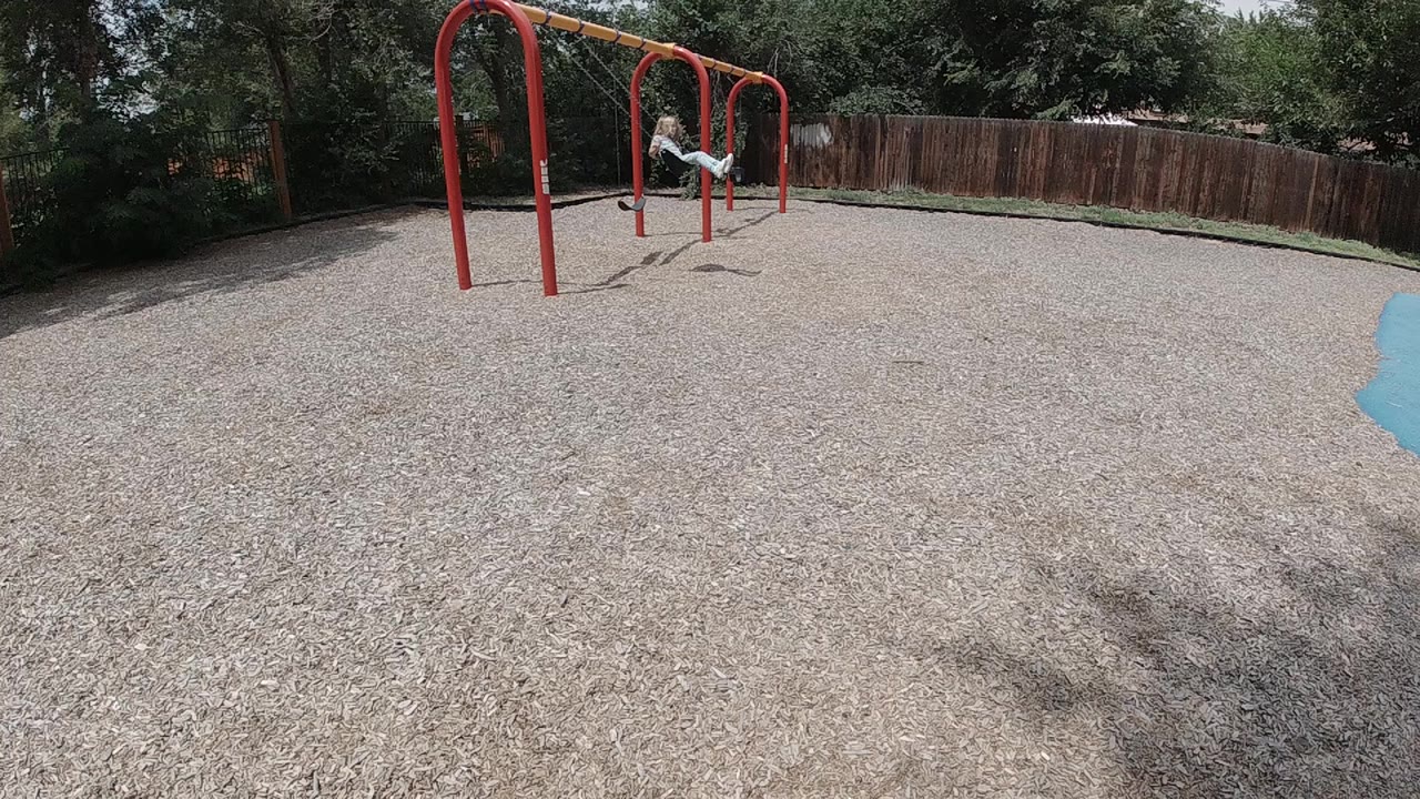 In Colorado, a child hangs near the first red monkey bar frame while pale gravel fills the foreground and a wooden fence lines the background.