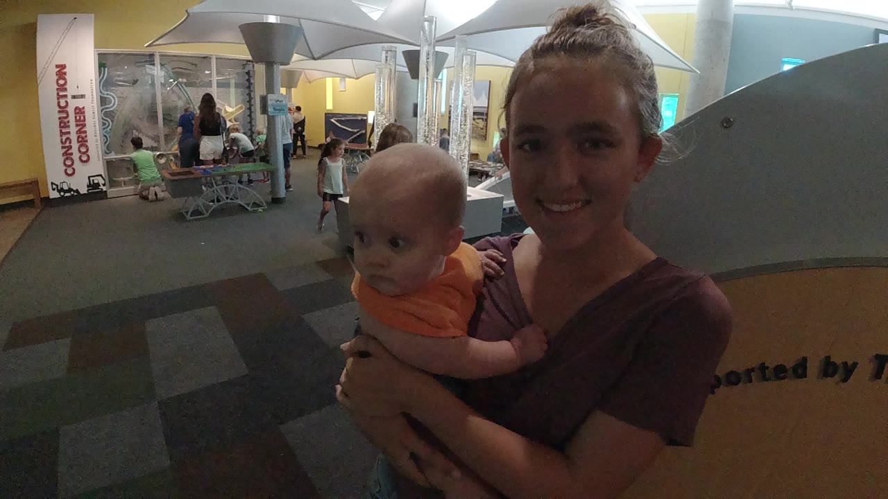 At the start of the Colorado clip, a smiling woman holds a baby in an orange shirt while event booths and seated attendees appear behind them.