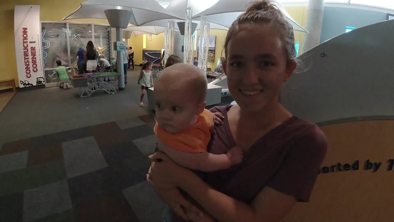 The baby looks away from the camera in the woman's arms, with a booth area, chairs, and distant visitors visible across the indoor venue.