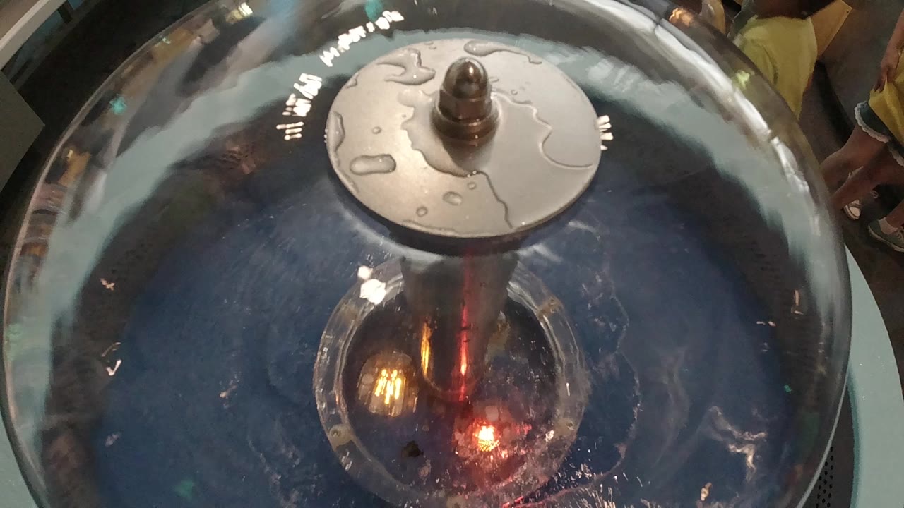 Overhead close-up of a large dark steel handpan filmed in Colorado, 2019; a lighter disc element sits on the instrument's crown; warm orange light reflects off the metallic dome at center; no visible people in frame.