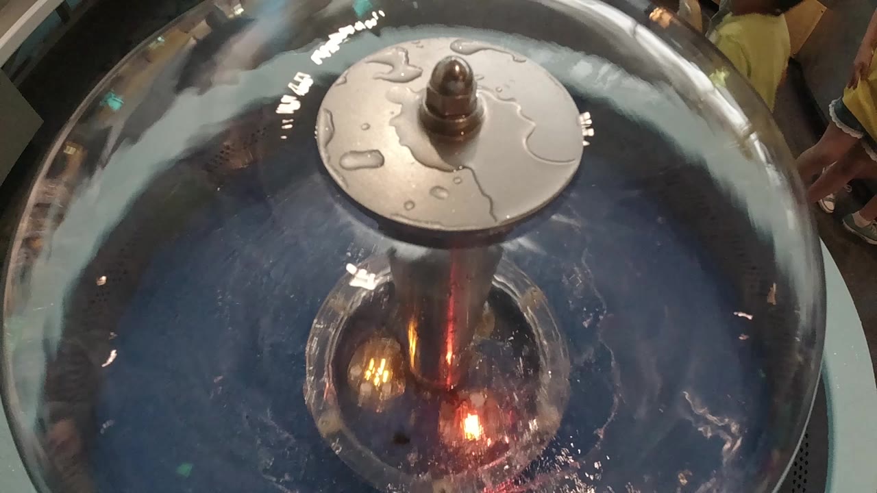 Overhead close-up of a dark steel handpan, 2019 Colorado; warm orange-red glow at the instrument's center suggests stage lighting reflecting off the spinning metallic dome; disc element on crown; no visible people in frame.