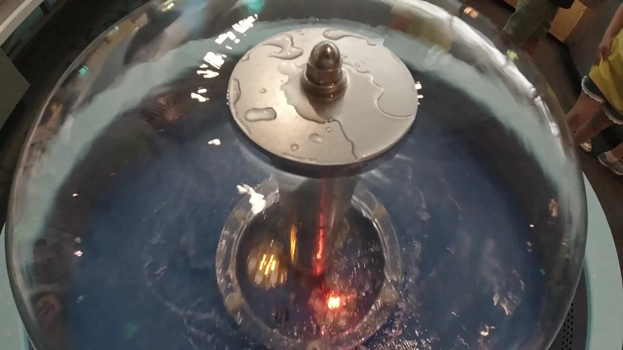 Overhead close-up of dark metallic handpan; central dome with disc element on crown; minimal warm reflection at center.