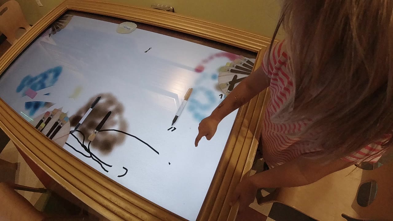 At 0:00, a child in a pink striped shirt stands at the right edge of a glowing wooden light table in Colorado, with black lines and blue and brown marks on the surface.