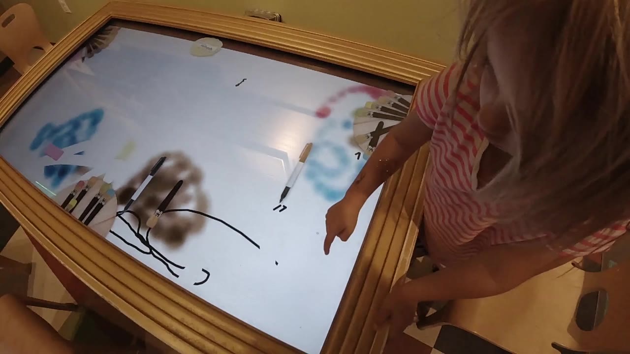 At 0:02, the child remains close to the right side of the wooden light table as abstract blue, brown, pink, and black markings are visible on the glowing panel.