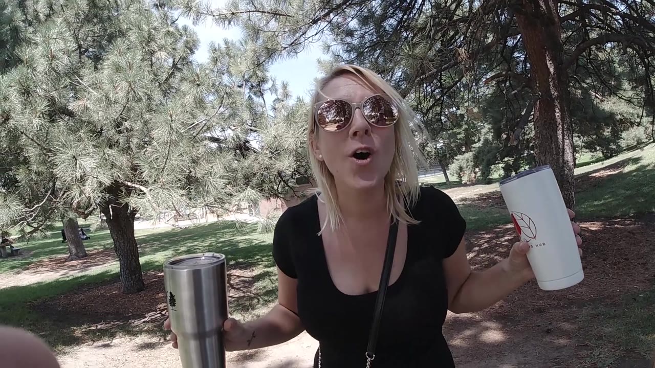The woman leans slightly closer to the camera in the Colorado park, holding the white travel cup beside her while trees fill the background.