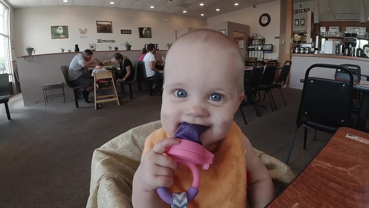 The baby faces the camera and bites the purple ice pop, with empty chairs, a wooden table edge, and seated cafe patrons visible behind in Colorado.