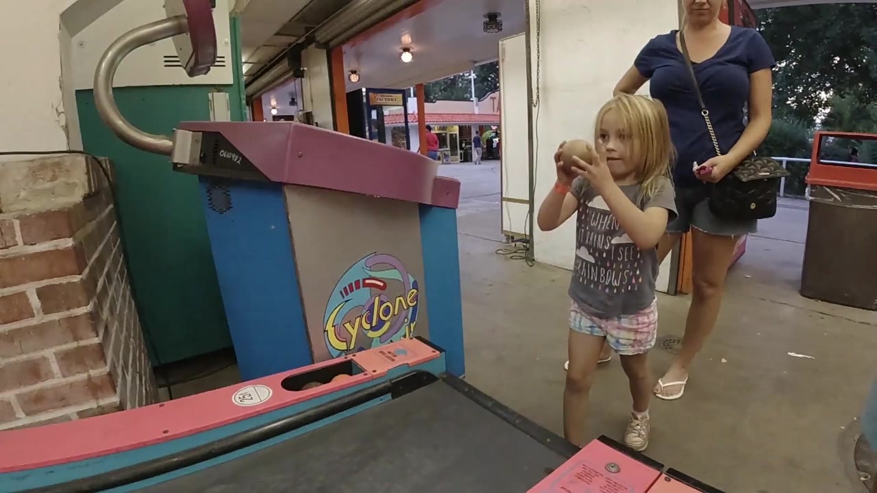 The child lifts the ball higher beside the colorful Cyclone game as the adult watches from the walkway behind her.