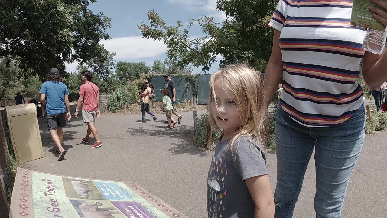In Colorado, a blonde young girl stands near a mounted park map while an adult in a striped shirt is cropped at the right and visitors walk along the path behind them.