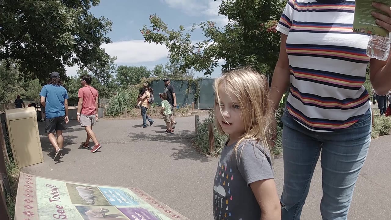 In Colorado, the child faces slightly toward camera beside the mounted map, with the adult cropped at the torso and people walking in the midground.