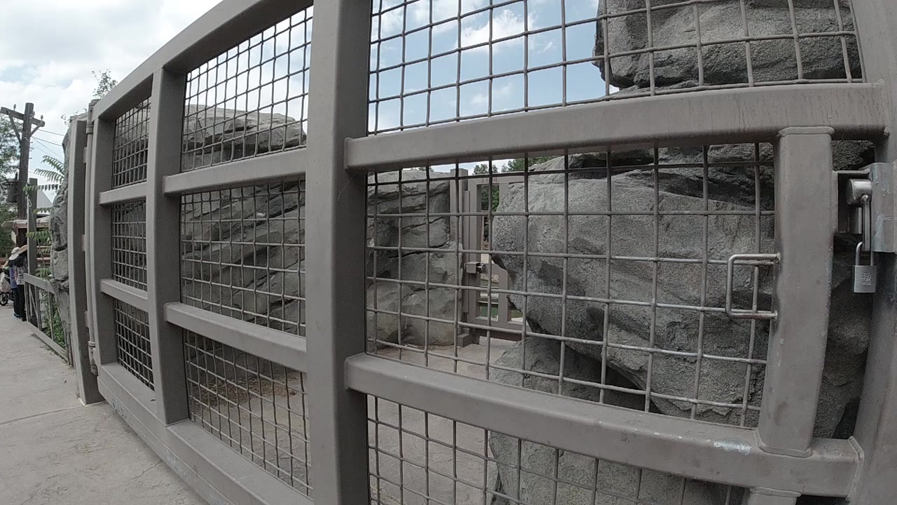 At the start of the Colorado clip, a grey metal mesh fence fills the foreground beside a concrete walkway, with rock-like surfaces behind it and no clearly visible people.