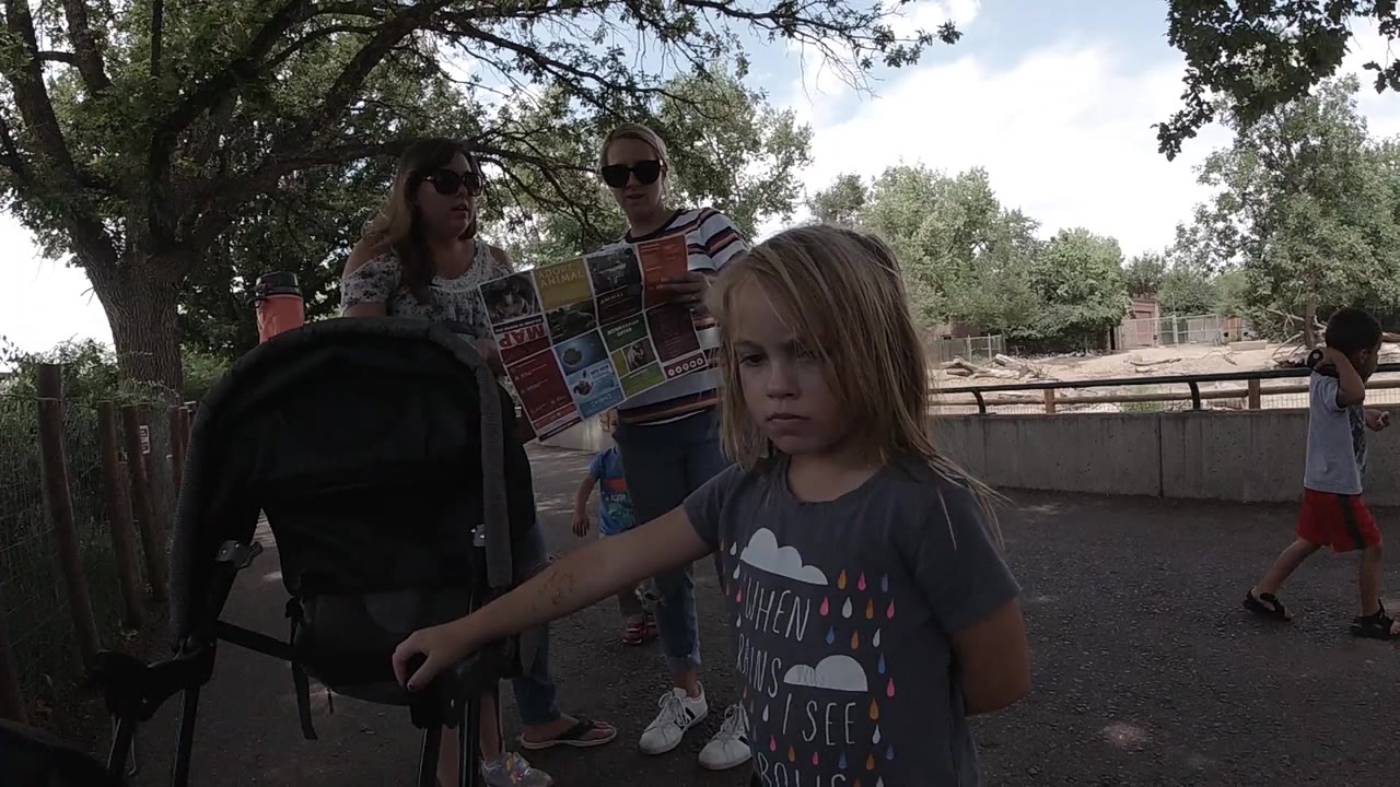In Colorado, a young girl stands beside a black stroller on a shaded paved path while two adults look at a colorful folded map behind her.