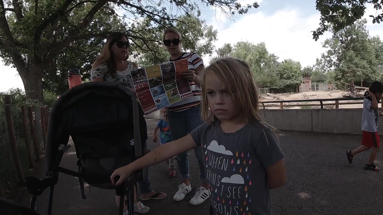 A child stands in front of a stroller while two adults with sunglasses and a map occupy the shaded midground beside a wooden fence in Colorado.