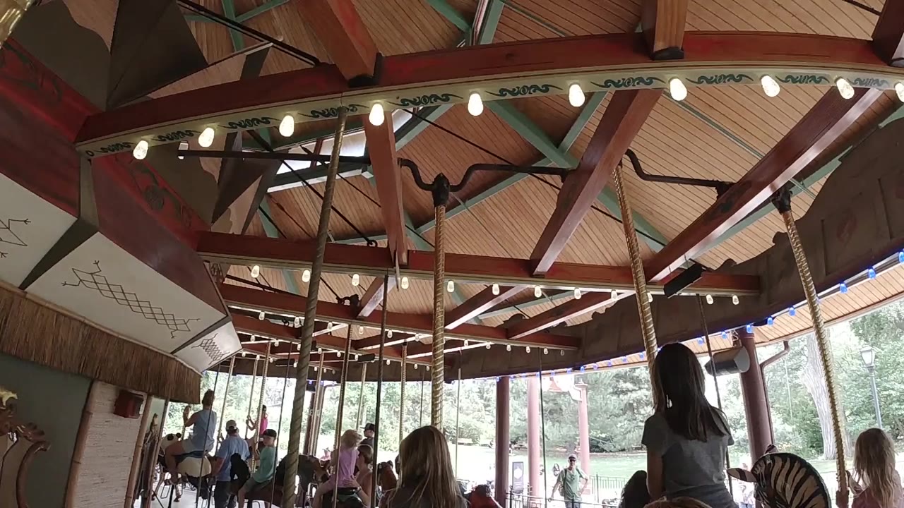 Colorado indoor carousel view at the start, showing wood ceiling panels, red beams, glowing bulbs, poles, windows, and people riding below.