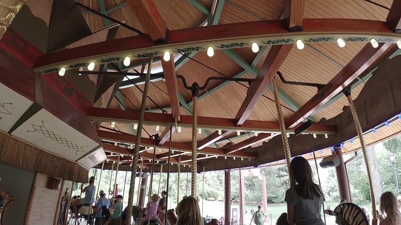 Colorado indoor carousel frame with the bulb-lit canopy across the top, a dark horse form near the beams, riders below, and daylight through tall windows.