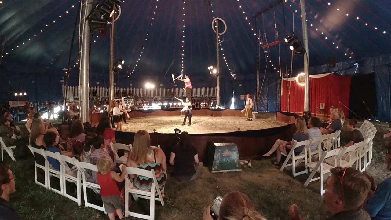 High-angle drone view of a circular circus ring lit by stage lights inside a tent in Colorado, 2019, with white folding chairs arranged around the ring and small figures present in the seating and performance area.