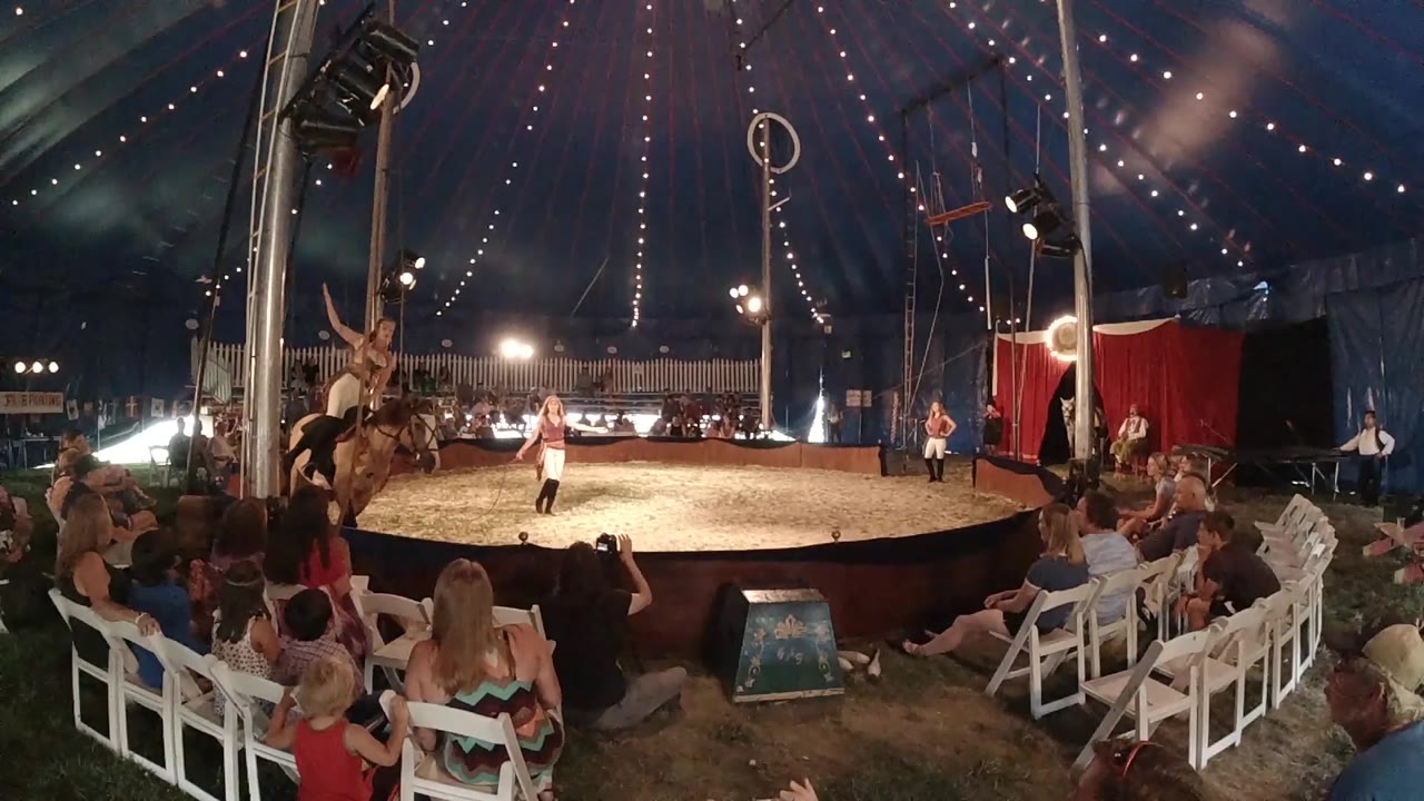 Wide view inside a Colorado circus tent as spectators watch a horse and rider near the left side of the dirt ring under string lights.