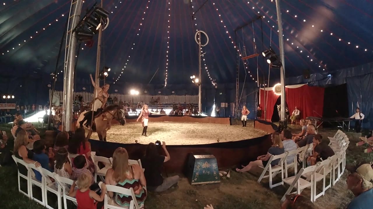 A performer stands on the horse as it moves through the left half of the Colorado circus ring, with seated spectators and overhead lights visible.