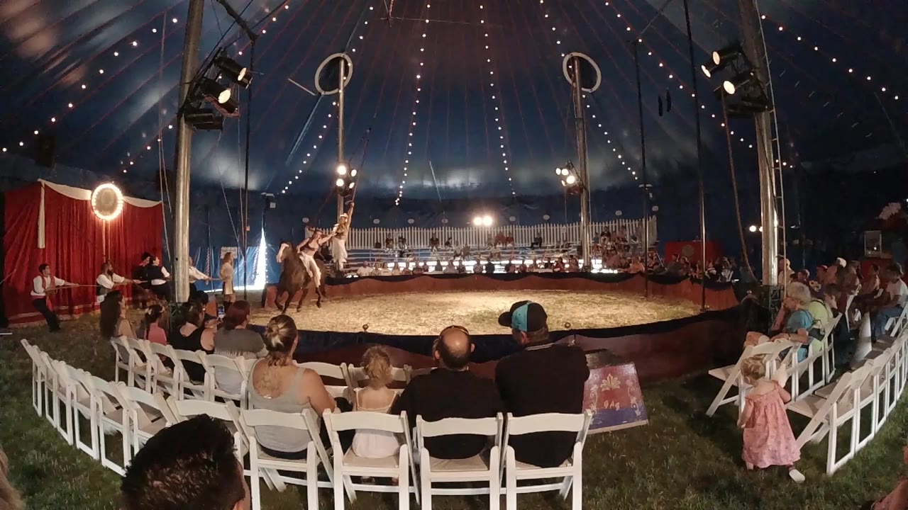 At the start of the Colorado circus clip, a rider on a dark horse circles the ring while an aerialist hangs above and spectators sit in white chairs.