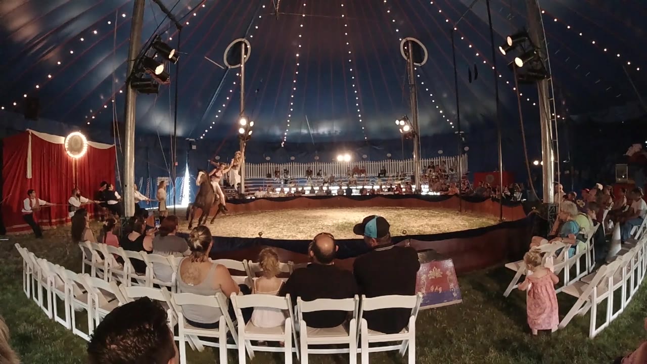 The rider continues around the sawdust ring as the aerialist hangs near center frame under rows of string lights in the Colorado tent.