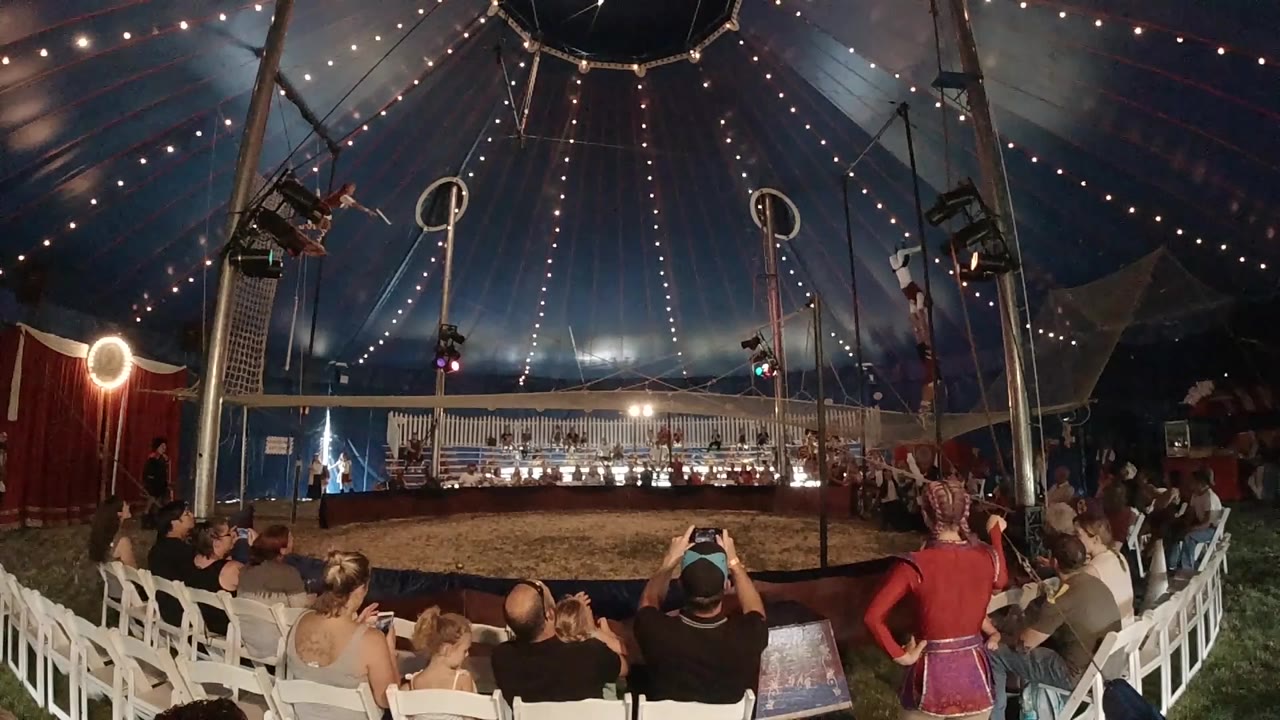 Wide view inside a Colorado circus tent shows seated spectators, white chairs, blue canopy lights, and aerial hoops hanging above the performance ring.