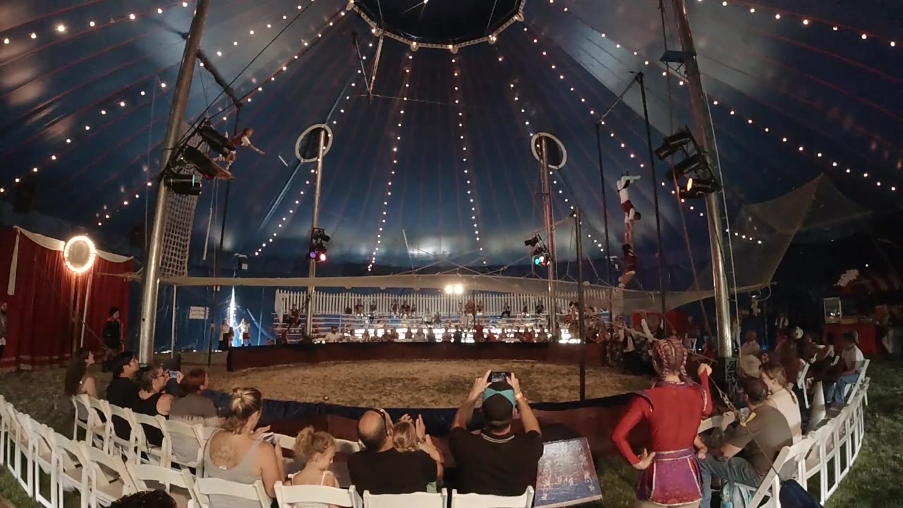 Spectators fill the foreground seating as two aerial rigs hang over the ring and stage lighting glows across the back of the Colorado tent.