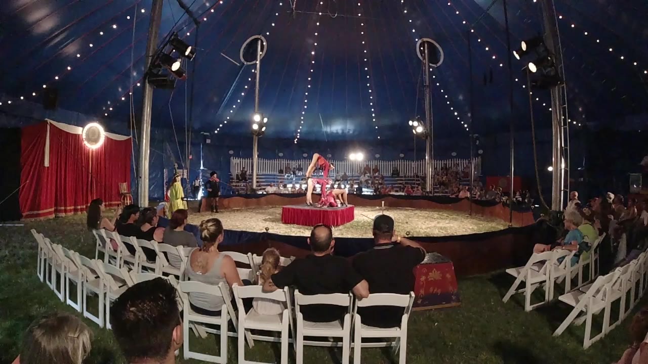 Opening frame of a Colorado circus tent performance, with white chairs in the foreground and a red-costumed performer centered under string lights.