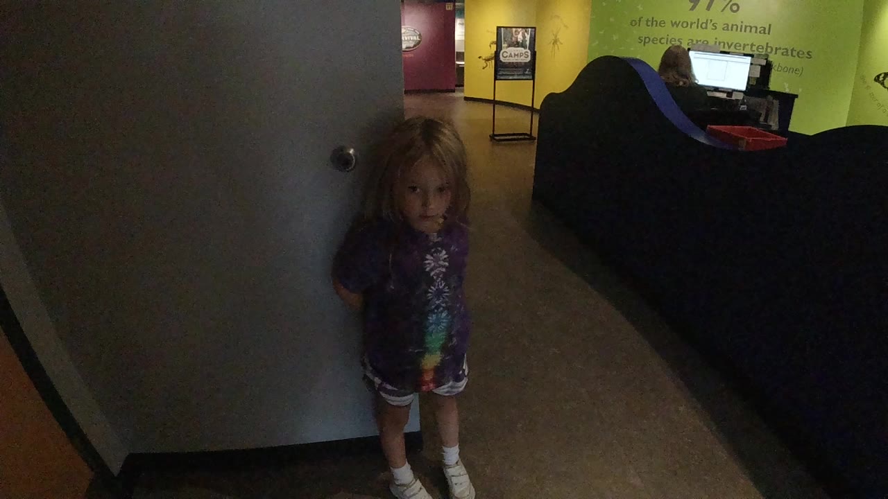 A toddler girl stands close to a light-colored interior door in Colorado, with a dark seating area and green wall visible behind her.