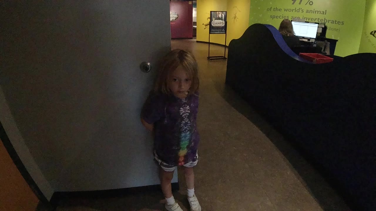 Child standing by door with hands near her sides.