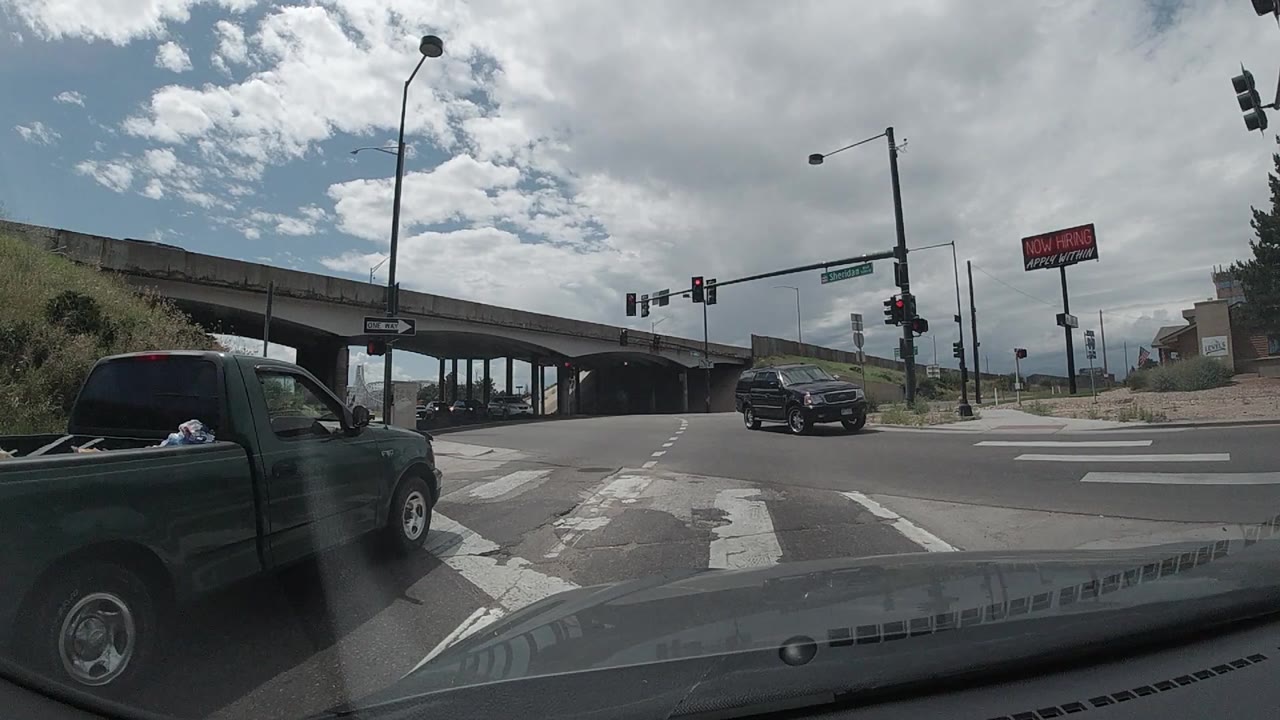 At 0:00, a Colorado dashcam view looks across a marked intersection under a concrete overpass, with a dark green pickup at left, a black SUV ahead, red traffic lights, and no visible people.
