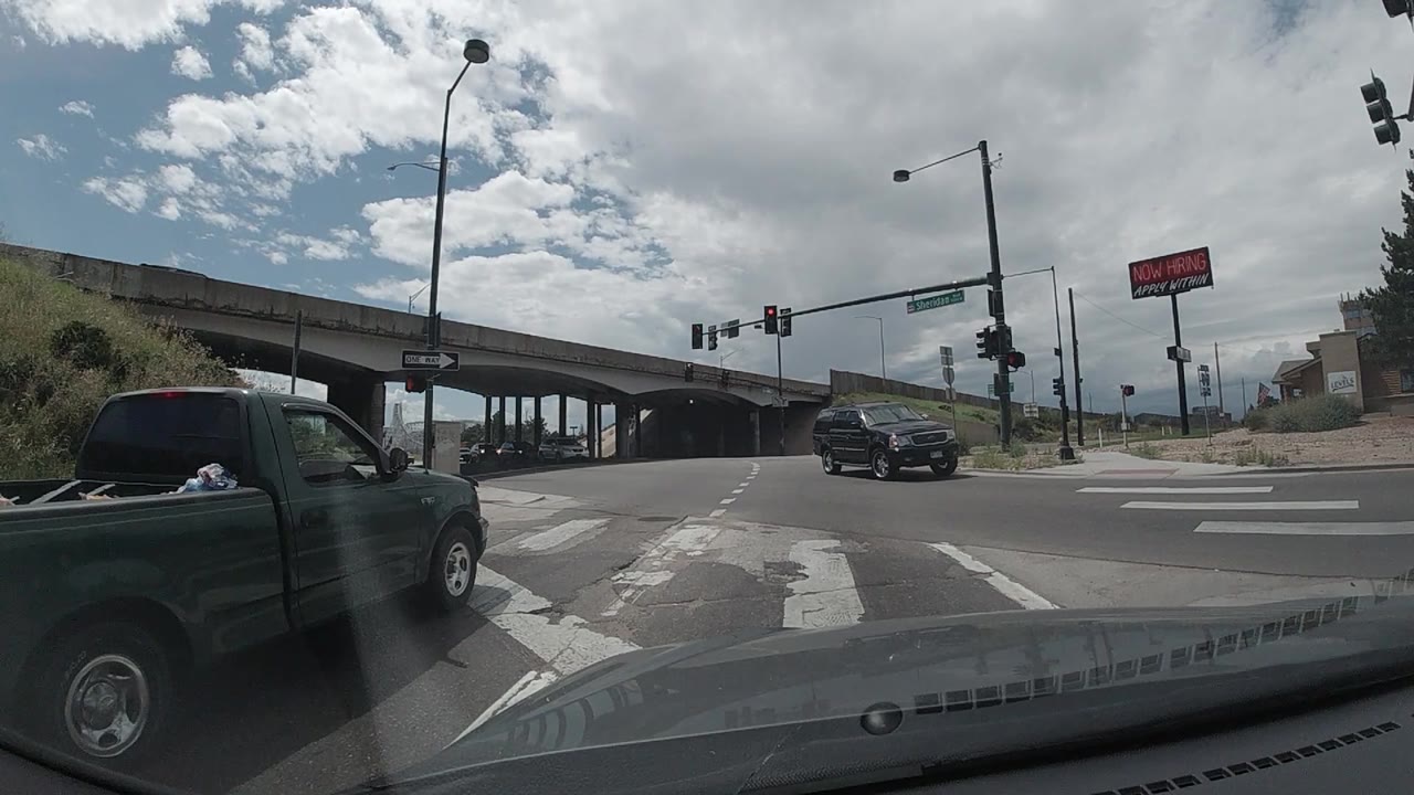 At 0:01, the windshield view holds on the Colorado intersection as the black SUV sits nearer the center lane area, the green pickup remains along the left edge, and overhead lights stay red with no visible people.
