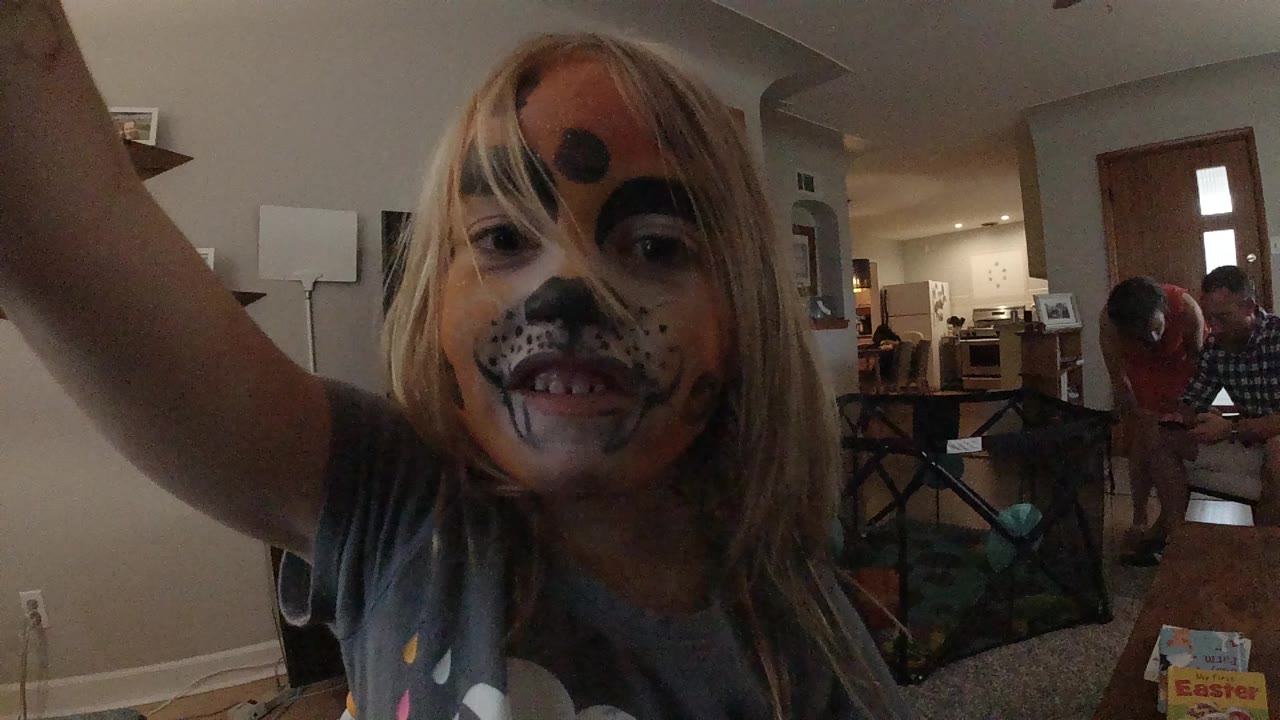 In a Colorado living room, a young girl with orange, black, and white face paint leans close to the camera while adults and a playpen sit in the background.
