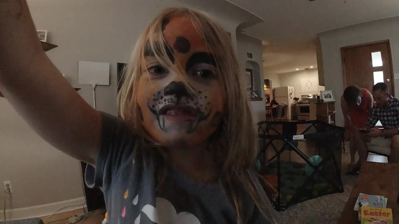 The child remains centered near the camera, with her raised arm at the edge of the frame and a home living area stretching behind her.