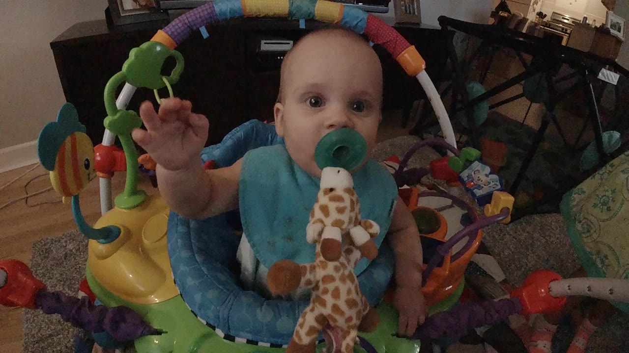 In Colorado, a baby with a green pacifier sits in a colorful activity center and raises one hand near the camera while a plush giraffe hangs at their chest.