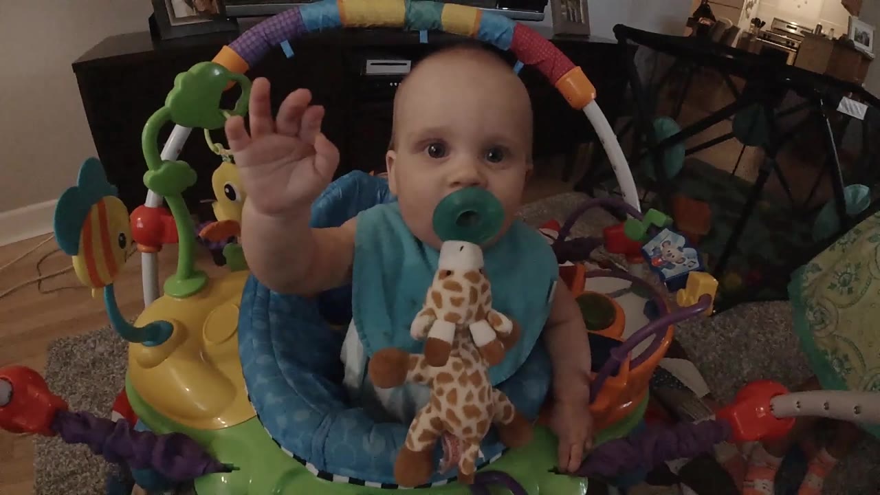 In Colorado, the baby raises an open hand beside their face while seated under the toy arch of a colorful activity center.