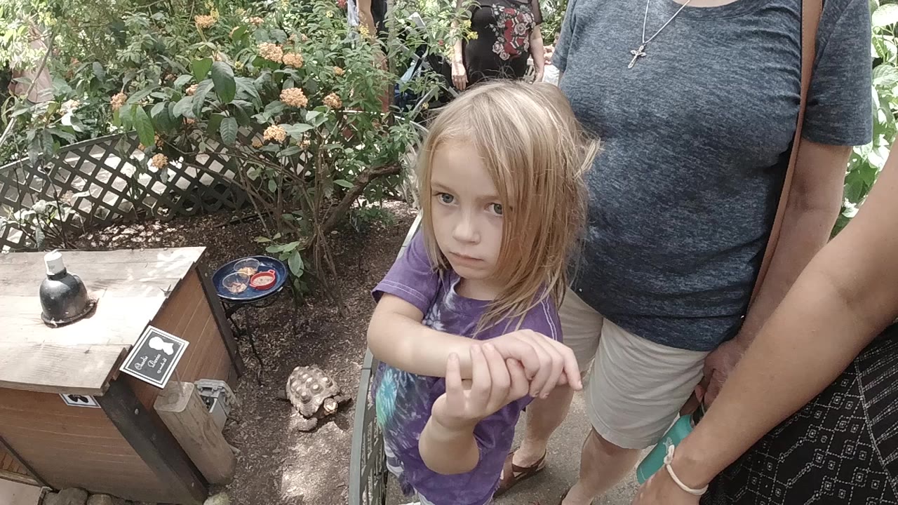 A young blonde girl in a purple shirt faces the camera beside adults in a Colorado garden with leafy plants, a lattice fence, and a small display sign nearby.