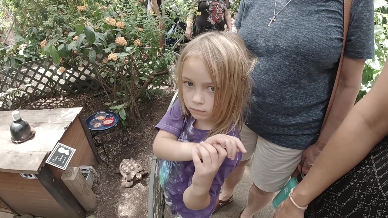 The young girl remains centered and looks directly toward the camera as adults stand close on both sides, with flowers, lattice, and a turtle-like ornament visible behind her.