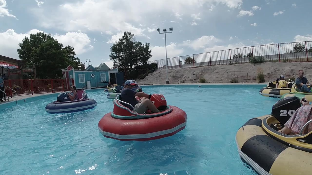 In Colorado, United States, riders sit in red and purple inflatable bumper boats on a turquoise pool, with a yellow boat edge in the foreground and fencing and trees behind them.
