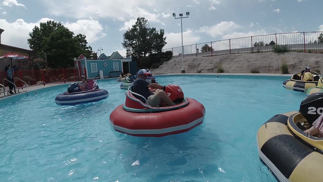 The red bumper boat drifts to the right side of the frame as a partial yellow boat enters the foreground, with blue structures and fencing still visible across the pool.