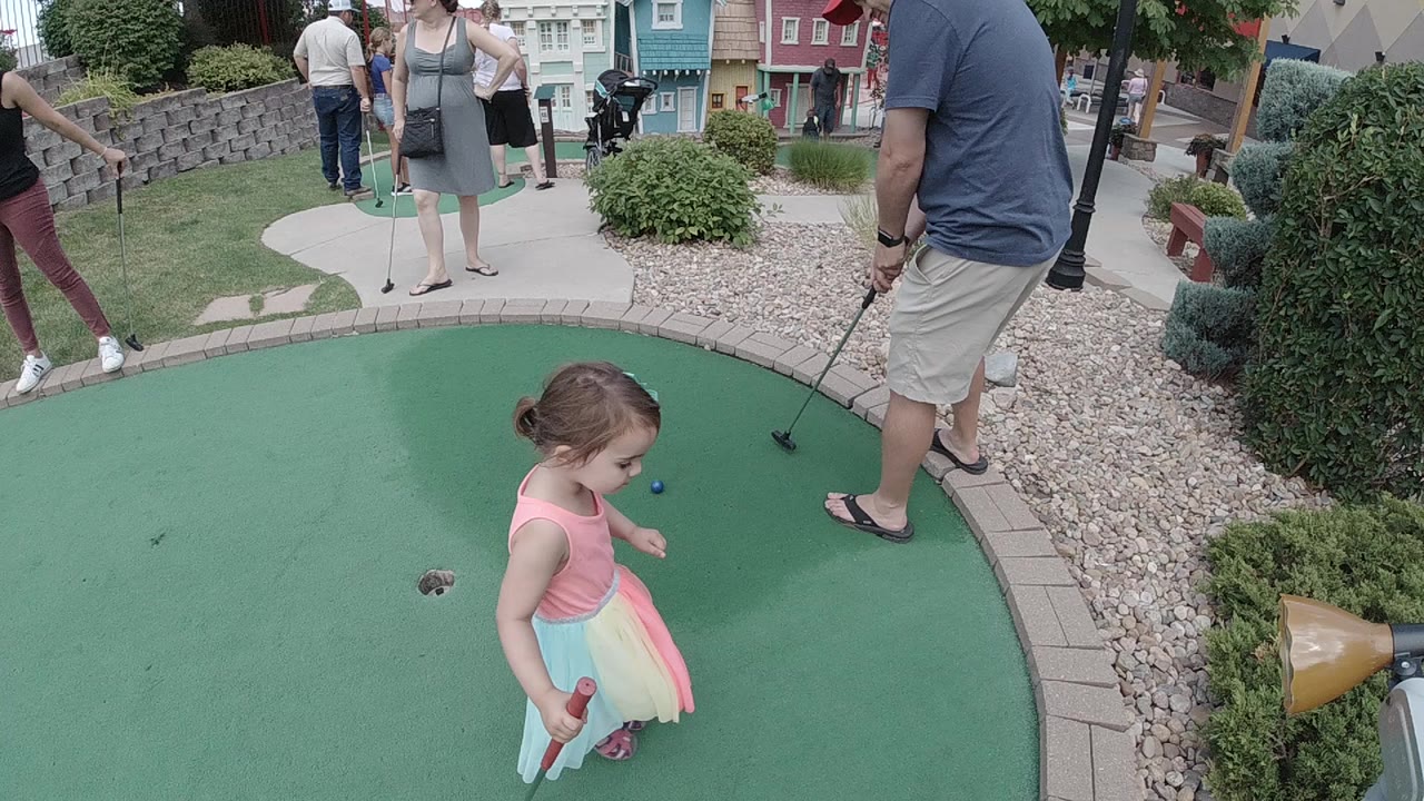 A young girl in a pastel dress stands on a Colorado miniature golf green while a man with a putter and other adults wait around the course edge.